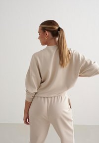 Set di abbigliamento loungewear beige con maglia oversize a spalle basse e pantaloni con vita elastica. Texture liscia, design minimale.