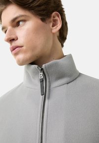 Lichtgrijze fleecejas met hoge kraag, metalen rits en logolabel. De stof heeft een zachte textuur en toont een minimalistisch ontwerp.