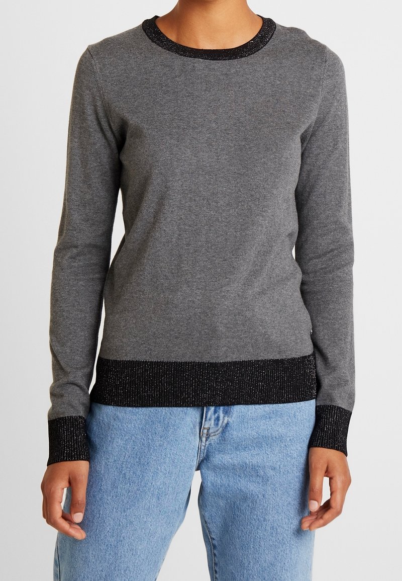 Personne portant un pull gris à manches longues avec col, poignets et ourlet côtelés noirs, assorti à un jean bleu clair, debout face à l’avant.