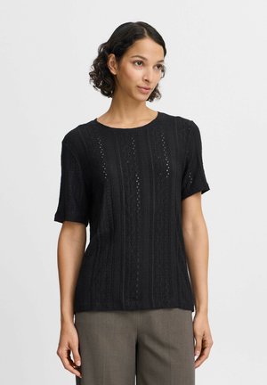 Schwarzes, kurzärmeliges Strickoberteil mit komplizierten Texturmuster und kleinen Öffnungen. Rundhalsausschnitt und lockere Passform, kombiniert mit grauen Hosen.