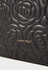 Juodas oda su siuvinėtais rožių raštais ir auksiniu "ANNA FIELD" logotipu, centriniu ant paviršiaus.