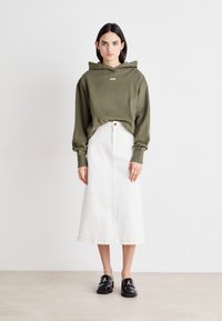 Felpa verde oliva con logo sul davanti, abbinata a una gonna midi in denim bianca e mocassini neri. Design a taglio dritto e vestibilità rilassata.