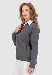 Felpa grigia con zip e una camicia bianca col collo sottostante, con tasca frontale e polsini a costine. Accessoriata con una sciarpa rossa a pois.
