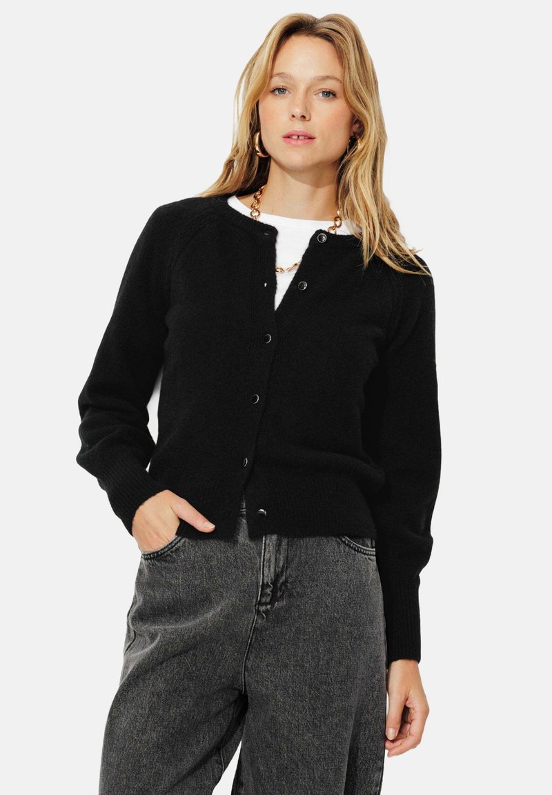 Cardigan noir à manches longues, avec des poignets côtelés et un col rond. Présente des boutons à l'avant et un tissu doux et texturé. Porté par-dessus un haut blanc.