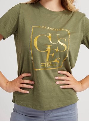 Frau trägt ein olivgrünes T-Shirt mit goldener Aufschrift "GUESS Los Angeles EST. 1981", die Hände in die Hüften gestützt, kombiniert mit hellblauen Jeans.