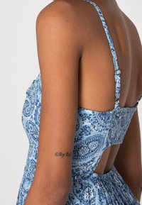 Modré šaty s paisley vzorem, tenkými ramínky, nařaseným dekoltem a otevřeným designem zad. Materiál vypadá lehký a strukturovaný.