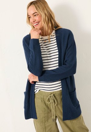 Lächelnde Frau, die eine marineblaue Strickjacke, ein weiß-schwarz gestreiftes Shirt und olivgrüne Kordelhose trägt und vor einer schlichten Wand steht.
