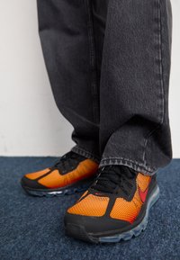 Černé a oranžové sportovní boty s vrchní částí z prodyšné síťoviny, černými tkaničkami a průhlednou podrážkou. Nošeny s tmavě šedými kalhotami na pozadí modrého koberce.