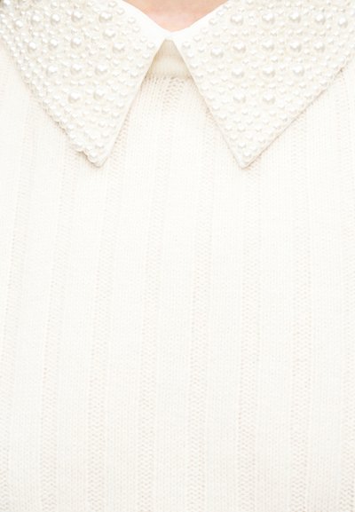 Vêtement en maille blanche avec un col pointu orné de perles régulièrement espacées, montrant le col et la partie supérieure de la poitrine.