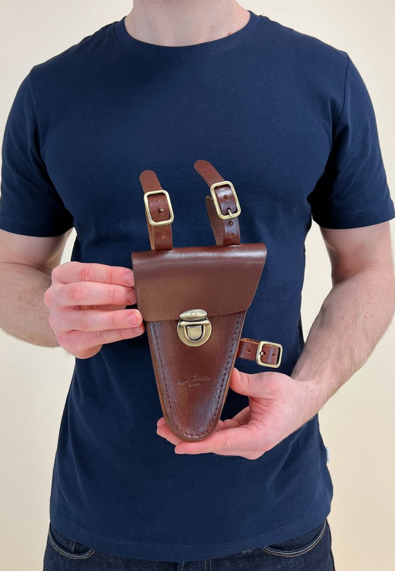 Bruine leren holster met een ronde onderkant, verstelbare bandjes en een metalen sluiting. Heeft een gladde afwerking en subtiele branding.