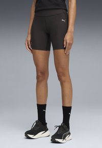 Γυναίκα που φοράει μαύρο σορτσάκι Puma, μαύρες κάλτσες Puma και μαύρα λευκά αθλητικά παπούτσια Puma, στέκεται σε γκρι φόντο.