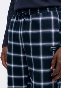 Tamno plave karirane flanel hlače s bijelim mrežastim uzorkom. Elastična pozadina s vezicom i oznakom branda na donjoj strani. Mekana tekstura.