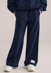 Tumši zili sporta bikses ar brīvu piegriezumu, sānos šuves un kabatas. Pievienotas gaišas krāsas kedas ar teksturētiem akcentiem.