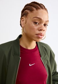 Alyvuogių žalia bombonešio striukė su sidabriniu užtrauktuku, dėvima virš burbulinės spalvos sportinės marškinėlių su mažuoju baltu "Nike" logotipu. Trumpi pinti plaukai.