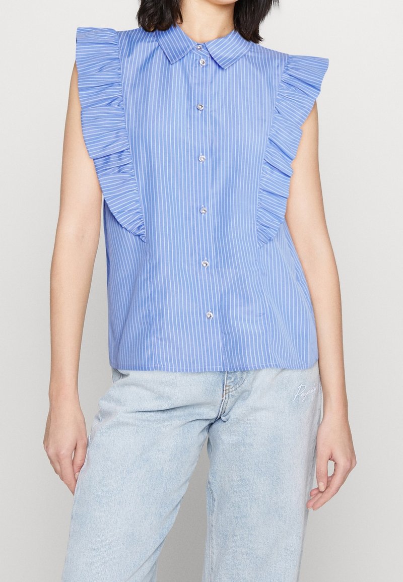 Camisetta senza maniche a righe blu con dettagli arricciati sulle spalle, chiusura con bottoni e vestibilità comoda, abbinata a jeans in denim blu chiaro.