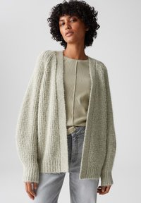 Lichtgroene cardigan met open voorkant, gebreide textuur, oversized mouwen en geribbelde boorden, gecombineerd met een fitted, lichtgroen top en denim jeans.