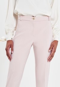Pantalons ajustés rose clair avec une texture lisse, dotés d'une taille bien ajustée et d'un détail en bouton doré à l'avant et aux poches latérales.