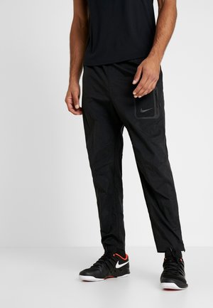 Mężczyzna ubrany w czarne spodnie sportowe z kieszenią na zamek oraz czarne buty Nike z białym logo i czerwonymi akcentami, stojący na białej podłodze.
