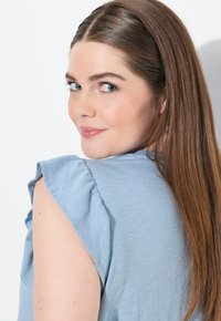 Top azul claro con mangas cortas y acampanadas. Tela suave, diseño simple y ajuste holgado. Cabello largo y castaño visible en el fondo.