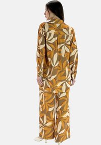 Completo a due pezzi floreale marrone, composto da una camicia a maniche lunghe e pantaloni a gamba larga, realizzato in un tessuto lucido con motivi di foglie in arancione e crema.