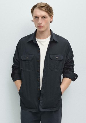 Junger Mann mit hellem Haar, der ein schwarzes Hemd über einem weißen T-Shirt trägt, mit den Händen in den Taschen stehend vor einem schlichten hellen Hintergrund.