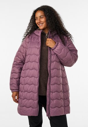 Kvinna med långt lockigt hår som ler, bär en lila vadderad jacka över en mörk tröja med dragkedja och svarta byxor, stående mot en vit bakgrund.
