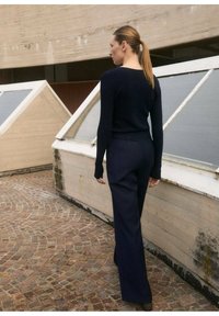Maglione a coste blu navy e pantaloni svasati su misura, entrambi realizzati in un tessuto testurizzato, mostrati su un pavimento con motivo di pietra e finestre.