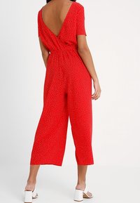 Röd jumpsuit med korta ärmar, öppen rygg, elastisk midja och vida ben; har vita prickar över hela. Bärs med vita sandaler.