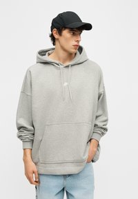 Νέος άνδρας με γκρι ανοιχτό φούτερ Nike, μαύρο καπέλο και ανοιχτό μπλε τζιν, κοιτάζει προς τα αριστερά με το ένα χέρι στην τσέπη.