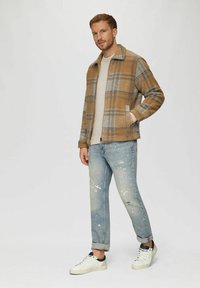 Kariertes Jackett in Braun- und Grautönen, mit Kragen, Vordertaschen und Reißverschluss, kombiniert mit hellblauen, distressed Jeans und weißen Sneakers.
