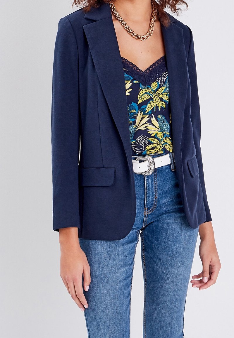 Femme portant un blazer bleu marine sur un haut fleuri orné de dentelle, jean bleu, ceinture blanche avec boucle décorative et collier chaîne doré.