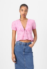 Rosa croppet blouse med V-hals, korte pufferm, knytfront og blonderdetaljer. Kombinert med et høyt liv denimbunad.