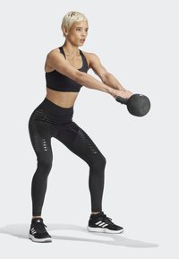 Czarny kettlebell, czarny stanik sportowy, czarne legginsy z wysokim stanem z błyszczącym wzorem w paski, czarne buty sportowe z białymi akcentami.
