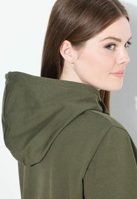 Una mujer con el cabello castaño lacio mira por encima del hombro, vistiendo una sudadera verde oliva con la capucha bajada, contra un fondo blanco.
