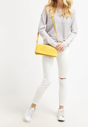 Žlutá semišová crossbody taška s minimalistickým obdélníkovým tvarem a stříbrnými detaily, nošená s světle šedým strukturovaným svetrem a bílými roztrhanými džínami.