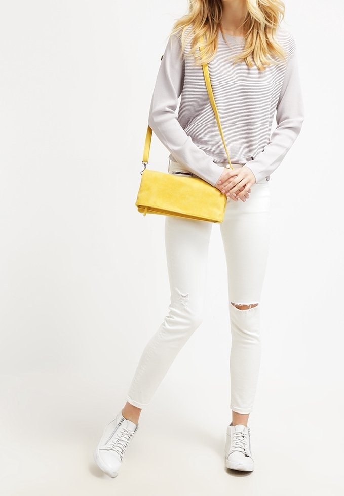 Žlutá semišová crossbody taška s minimalistickým obdélníkovým tvarem a stříbrnými detaily, nošená s světle šedým strukturovaným svetrem a bílými roztrhanými džínami.