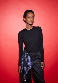  schwarzes Langarmshirt, kombiniert mit navyfarbener Hose. Ein kariertes Hemd in Schwarz und Lila ist um die Taille gebunden. Der Hintergrund ist rot.