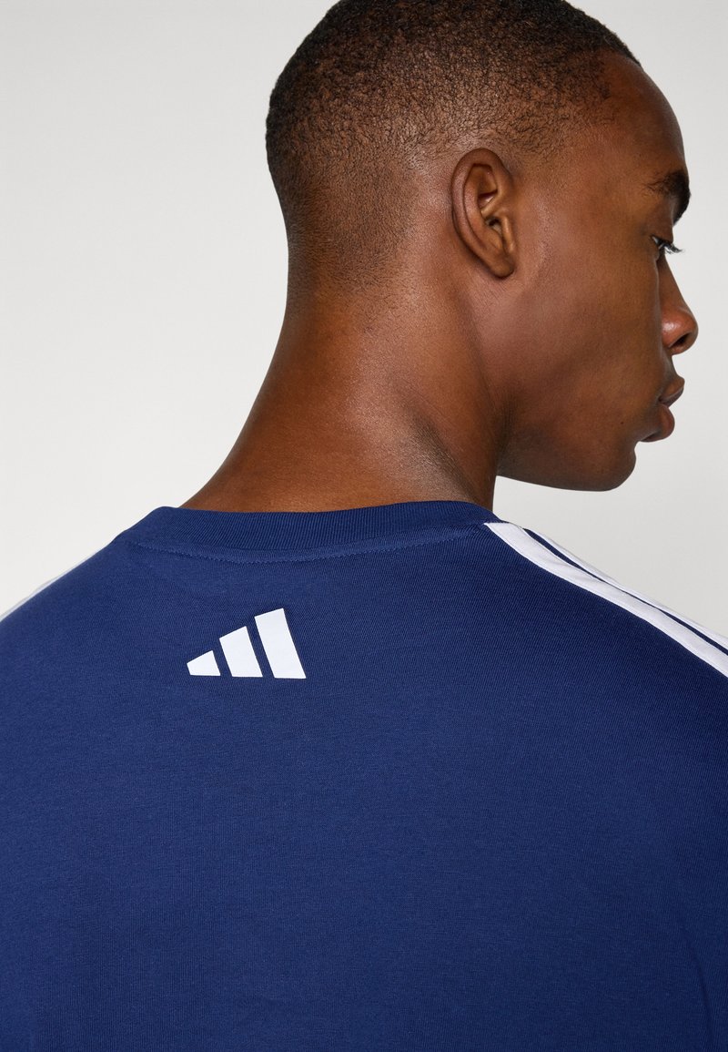 Granatowa bawełniana koszulka z białym logo Adidas na plecach, z okrągłym dekoltem i kontrastowymi paskami na ramionach.
