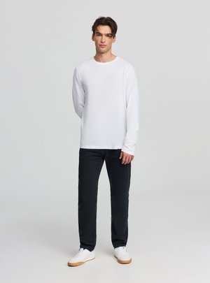 Maglietta bianca a maniche lunghe in cotone, scollo rotondo, abbinata a jeans neri e sneakers bianche con suola color cuoio. Design semplice con dettagli minimi.