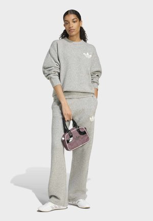 Femme en sweat-shirt Adidas gris et pantalon, tenant un petit sac à main violet avec des poignées noires, portant des baskets blanches, debout sur un fond uni.