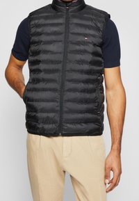 Czarna pikowana kamizelka z wysokim kołnierzem, zapięciem na zamek, bocznymi kieszeniami i poziomymi przeszyciami, w zestawieniu z granatową koszulą z krótkim rękawem.