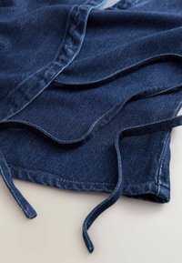 Tummansininen denimmateriaali, jossa on kaarevat reunat ja nyörit. Pinta on sileä, ja saumoissa näkyy ompelun yksityiskohtia.