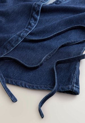 Jeansowa tkanina w głębokim granatowym kolorze, z zaokrąglonymi krawędziami i sznurkami. Tekstura jest gładka z widocznymi detalami przeszyć wzdłuż szwów.