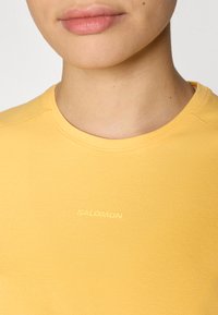 T-shirt en coton jaune avec un col rond, arborant un logo "SALOMON" subtil en contraste. Texture lisse et détails de design simples.