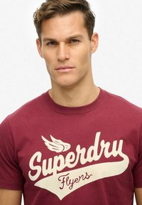 Bordowa bawełniana koszulka z dużym kremowym napisem "Superdry Flyers" oraz elementem graficznym skrzydeł. Okrągły dekolt, krótkie rękawy, gładka tekstura.