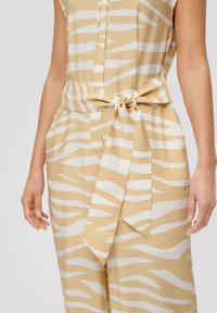 Beige Jumpsuit mit weißen Tigerstreifen, leichtes Material, kurze Ärmel, tailliert mit Gürtel und Seitentaschen. Strukturierte Optik mit Schleifen-Detail.