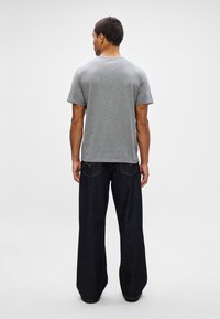 Mężczyzna odwrócony tyłem, ubrany w prostą szaro-skalową koszulę z krótkim rękawem oraz luźne, ciemnoniebieskie dżinsy, na tle białym.