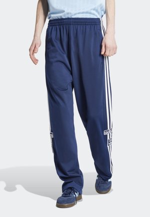 Mandlig model iført marineblå Adidas træningsbukser med hvide striber ned langs siderne og marineblå sneakers, stående på en hvid baggrund.