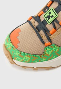 Bézs színű sportcipő texturált felsőrésszel, narancs és zöld részletekkel, geometriai mintával a talpon és fekete fűzőkkel.