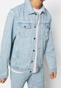 Light blue denim jacket with embossed paisley patterns, decorative stitching, two front pockets, and button closures. Worn over a white shirt.